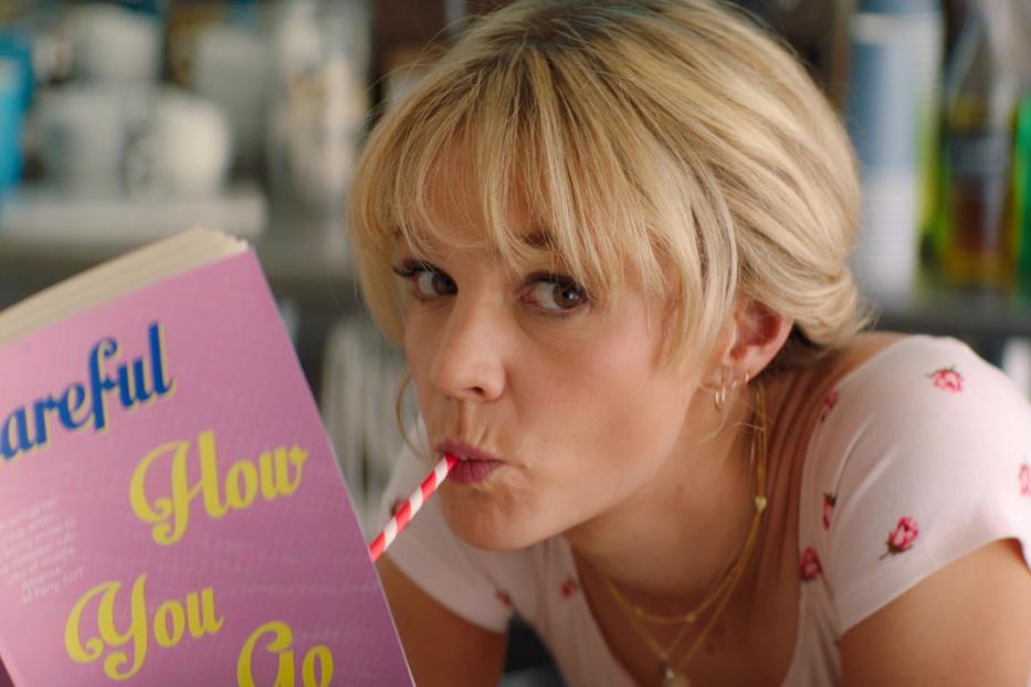 Eine blonde Frau trinkt mit einem Strohhalm und liest das Buch „Careful How You Go“.