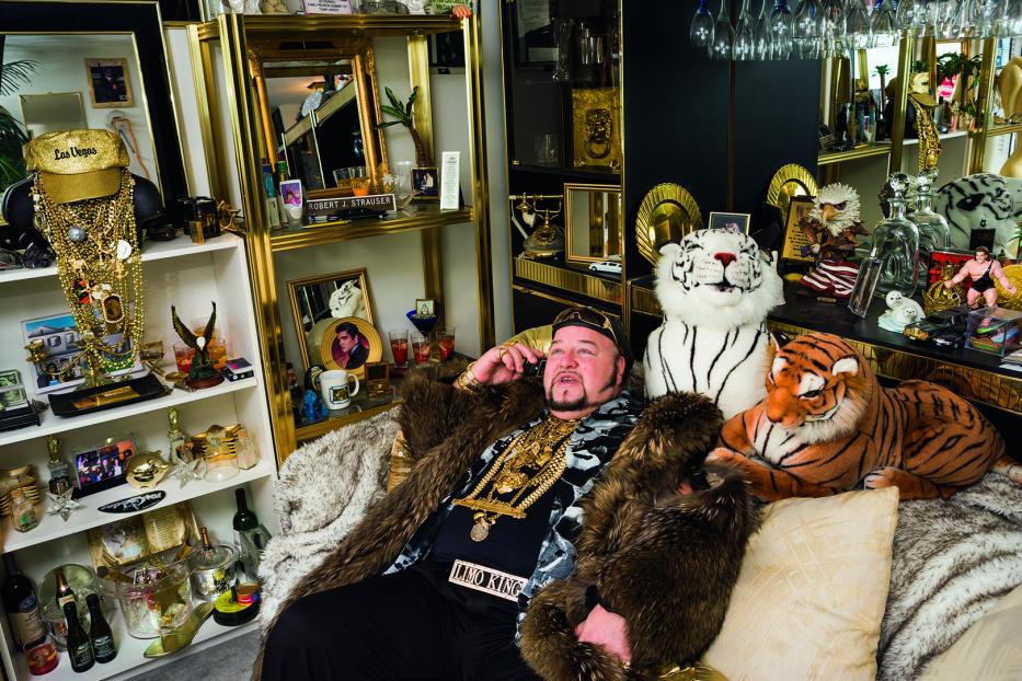 Ein Mann, vermutlich „Limo King“, sitzt mit Pelzmantel und viel Goldschmuck auf einem Sofa und telefoniert.