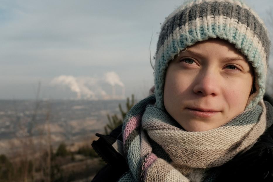 Greta Thunberg mit Mütze und Schal vor einer verschmutzten Landschaft.
