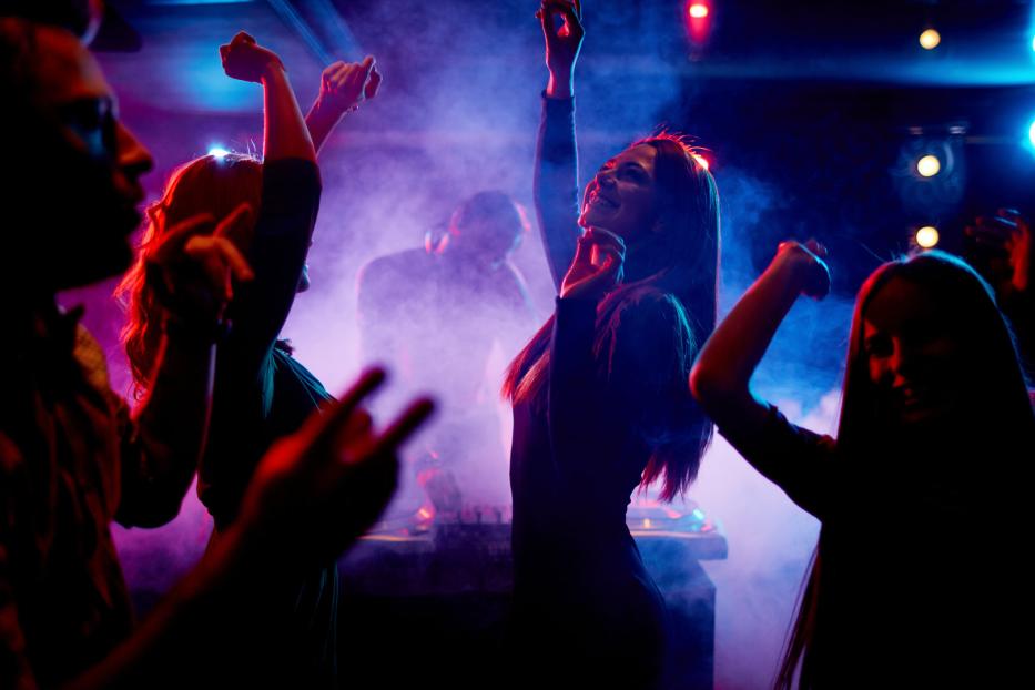 Menschen tanzen in einem Nachtclub mit farbiger Beleuchtung und einem DJ im Hintergrund.