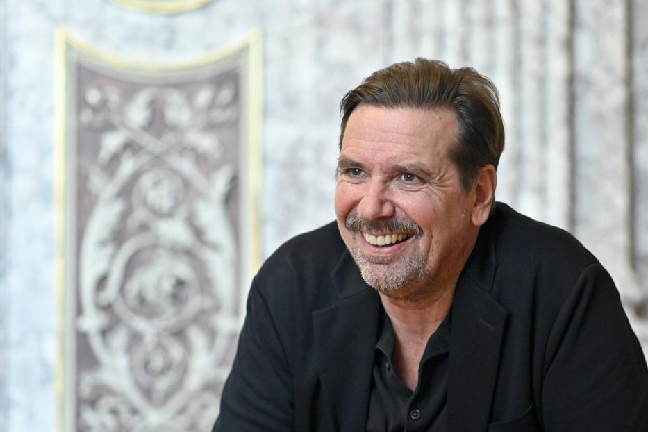 Ein lächelnder Mann mit Schnurrbart und dunklem Sakko vor einer verzierten Wand.
