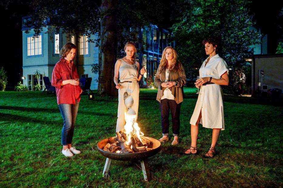 Vier Frauen stehen abends mit Getränken um eine Feuerschale im Garten.