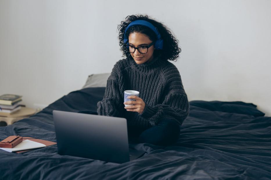 Eine Frau sitzt mit Kopfhörern auf dem Bett, arbeitet am Laptop und hält eine Tasse in der Hand.