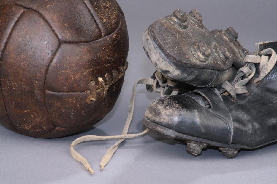 Ein alter, abgenutzter Fußball und ein Paar Fußballschuhe stehen auf einem grauen Untergrund.