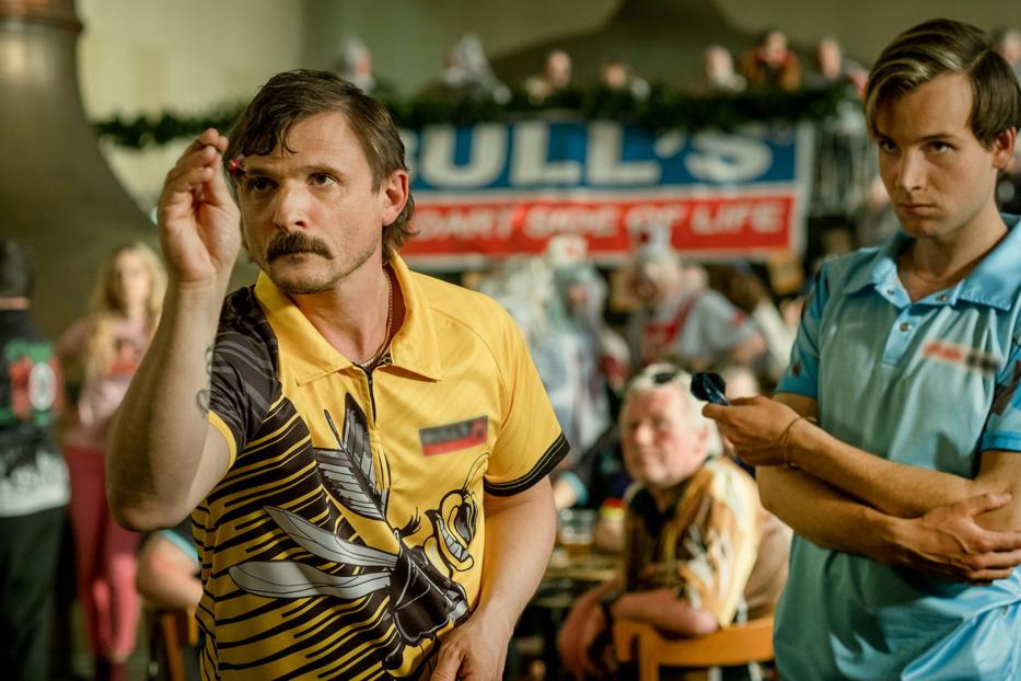 Ein Mann mit Schnurrbart zielt mit einem Dartpfeil auf eine Dartscheibe.