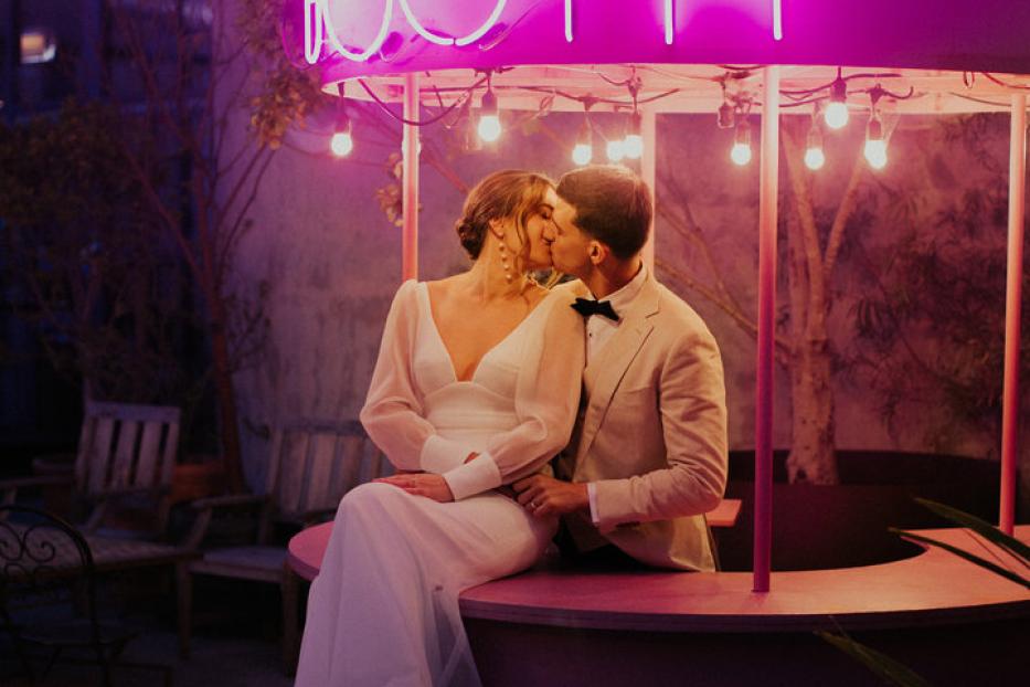 Ein Brautpaar küsst sich vor einem rosa „Kissing Booth“ mit Neonbeleuchtung.