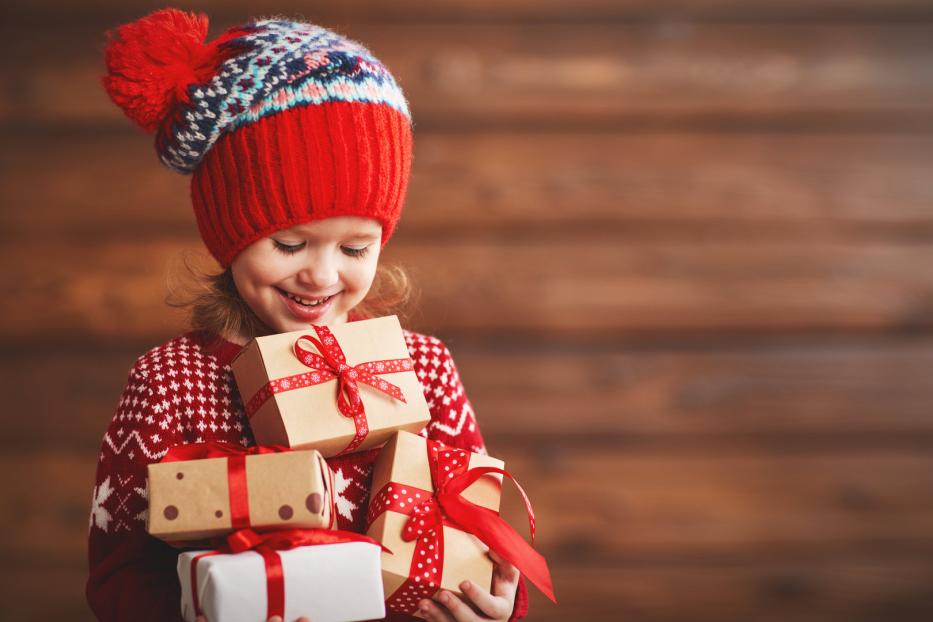 Ein lächelndes Mädchen mit Mütze hält mehrere Weihnachtsgeschenke im Arm.