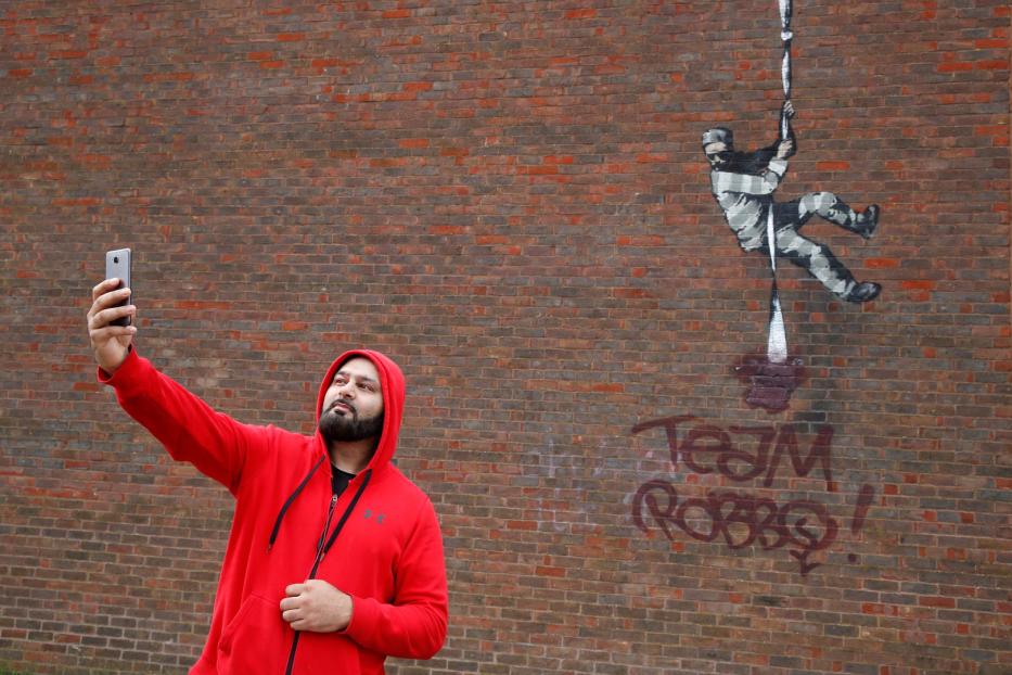 Ein Mann macht ein Selfie vor einer Backsteinmauer mit einem Banksy-Graffiti.