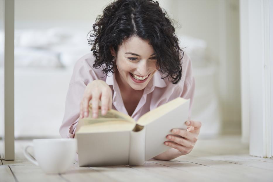 Eine lachende Frau liest ein Buch, während eine Tasse neben ihr auf dem Boden steht.