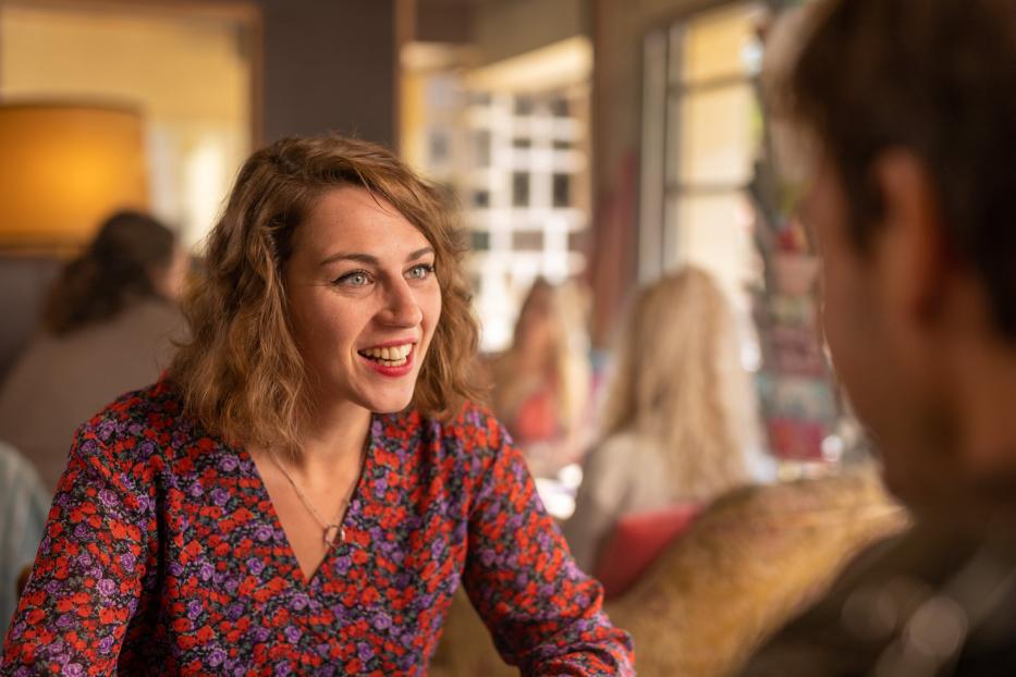 Eine lächelnde Frau mit blumigem Oberteil sitzt in einem Café und unterhält sich.