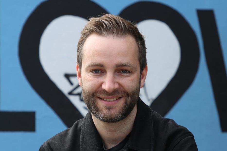 Ein Mann mit Bart lächelt vor einer Wand mit einem Herz-Graffiti.