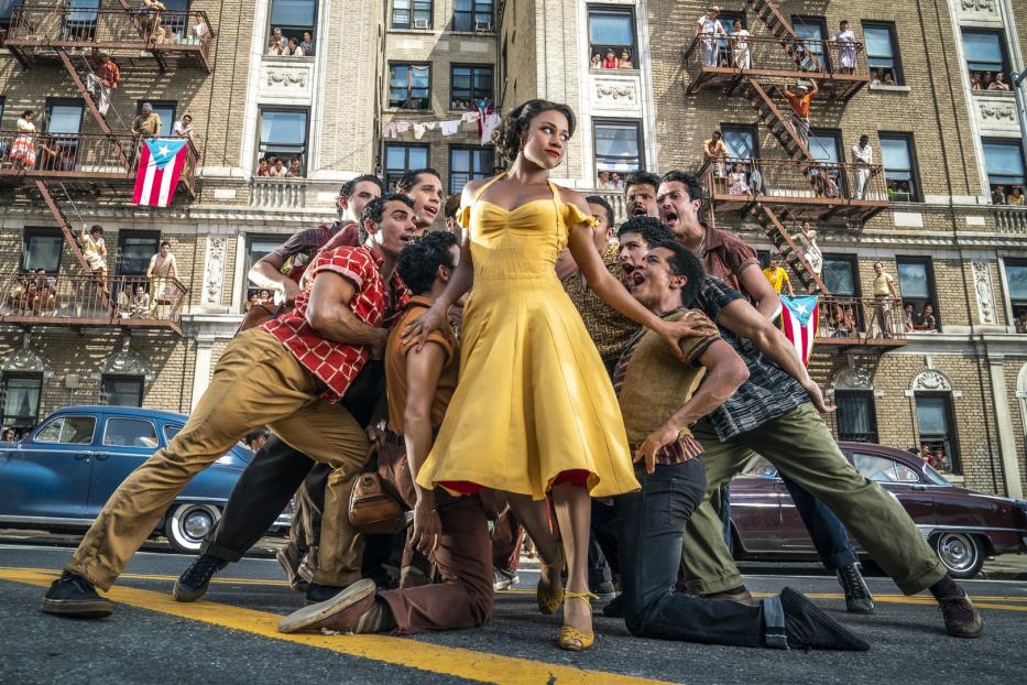 Eine Frau in einem gelben Kleid steht inmitten einer Gruppe von Tänzern vor einem Backsteingebäude mit puertoricanischen Flaggen.