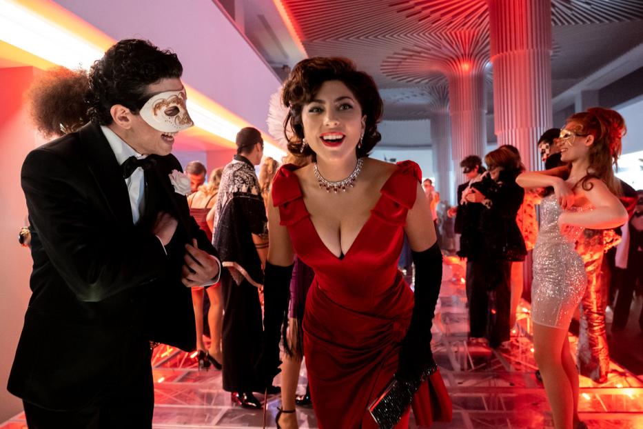 Lady Gaga, in einem roten Kleid, lächelt auf einer Maskerade mit anderen Gästen.