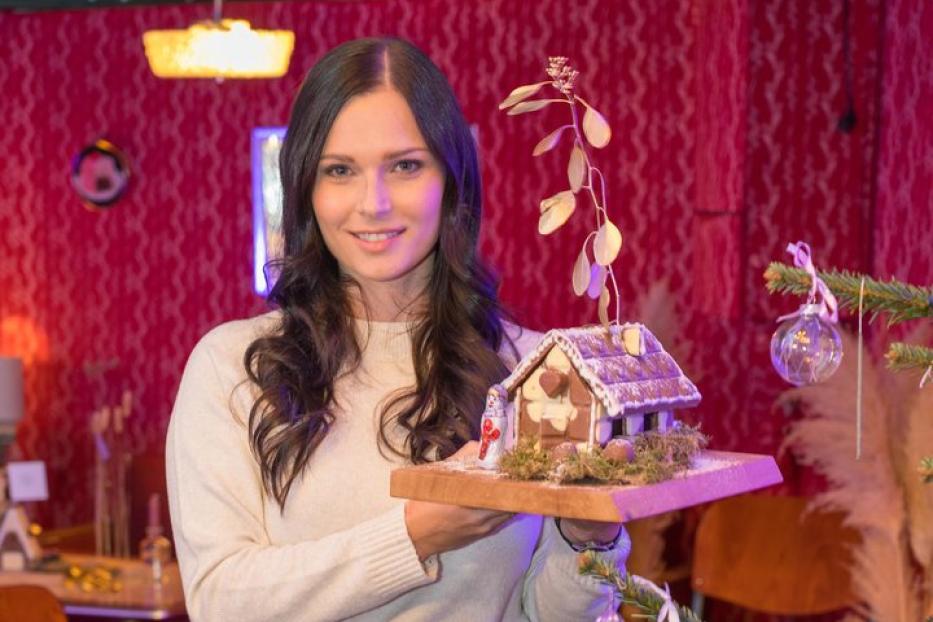 Eine Frau hält ein verziertes Lebkuchenhaus vor einem Weihnachtsbaum.