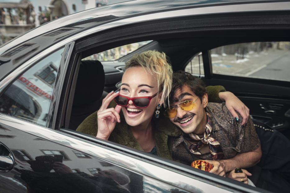 Eine lachende Frau mit Sonnenbrille und ein Mann mit gelber Brille sitzen in einem Auto.