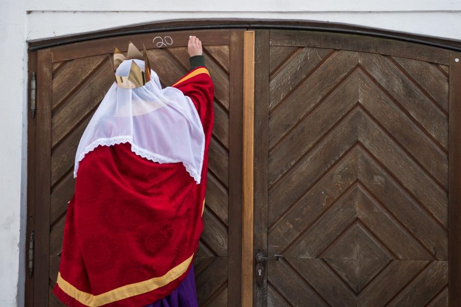 Ein als Sternsinger verkleidetes Kind schreibt mit Kreide „20“ an eine Holztür.