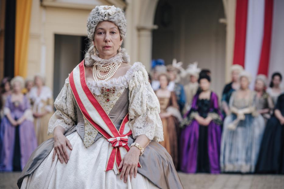 Eine Frau in historischer Kleidung mit Perlenkette und weiß-roter Schärpe vor einer Gruppe von Menschen.