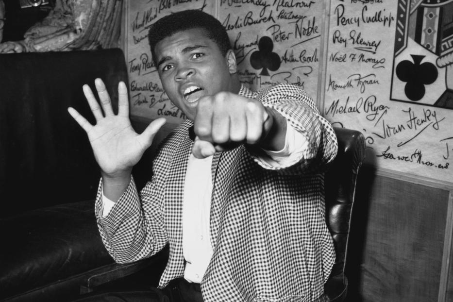 Muhammad Ali posiert mit erhobener Faust und offener Hand vor einer Wand mit Unterschriften.