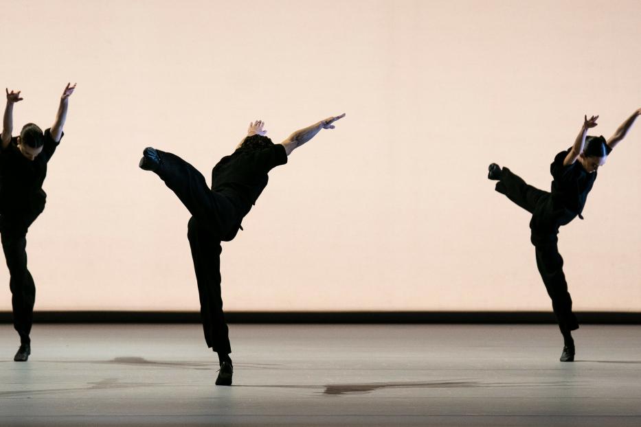 Drei Tänzer in schwarzen Kostümen führen eine synchrone Tanzroutine auf einer Bühne auf.