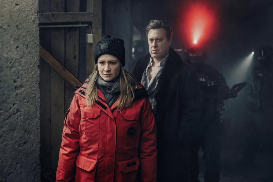 Eine Frau in roter Jacke und ein Mann stehen vor einem SWAT-Team mit rotem Licht.