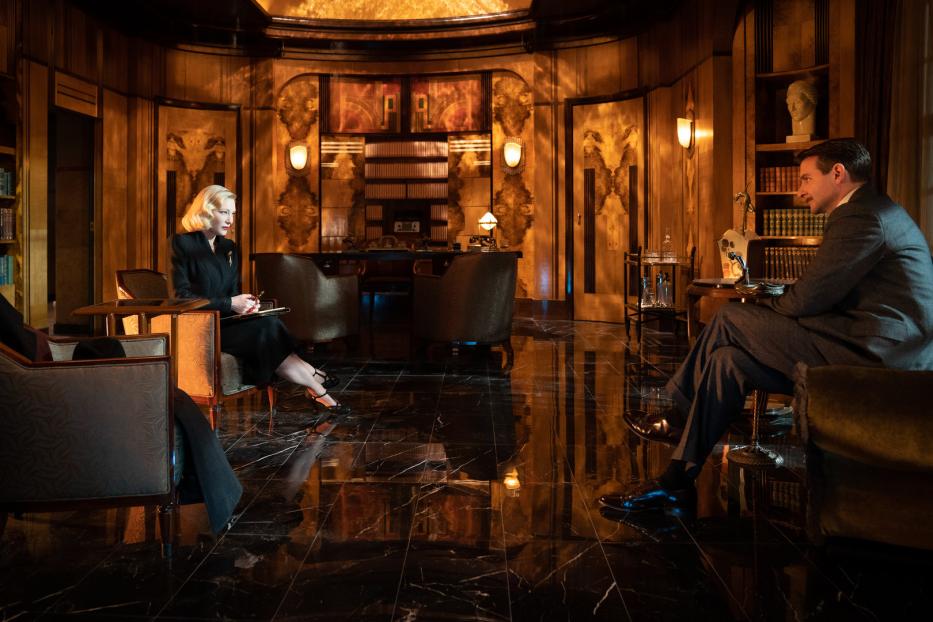 Cate Blanchett und Matt Bomer sitzen sich in einer eleganten Büroszene gegenüber.