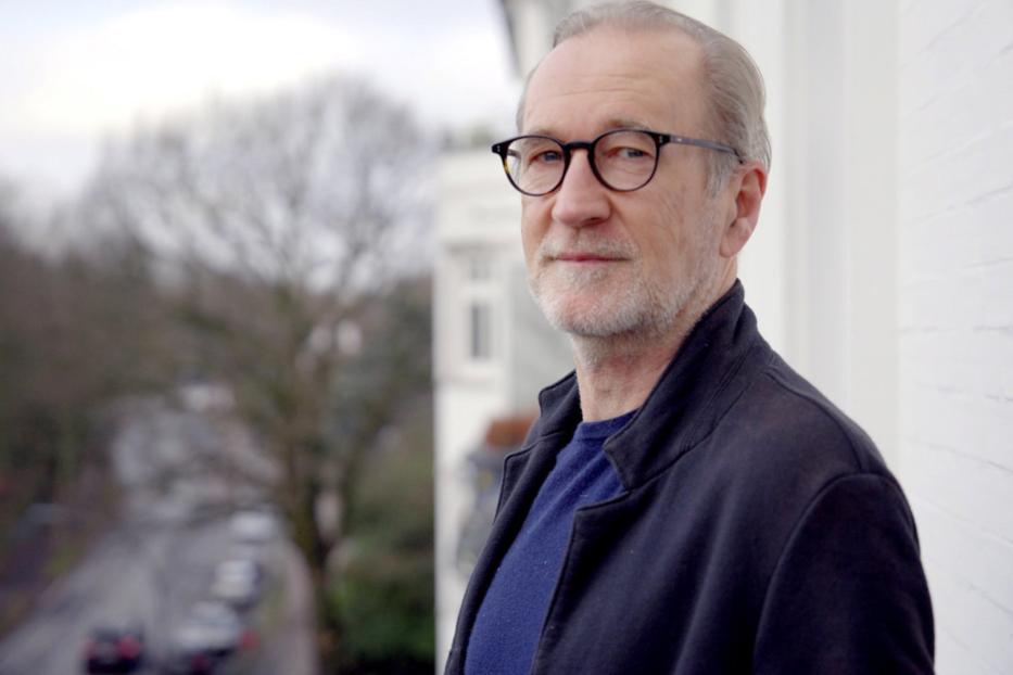 Ein Mann mit Brille steht auf einem Balkon und blickt in die Ferne.