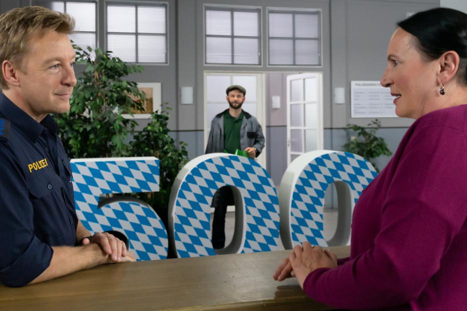 Ein Polizist spricht mit einer Frau vor großen, blau-weiß karierten Buchstaben.