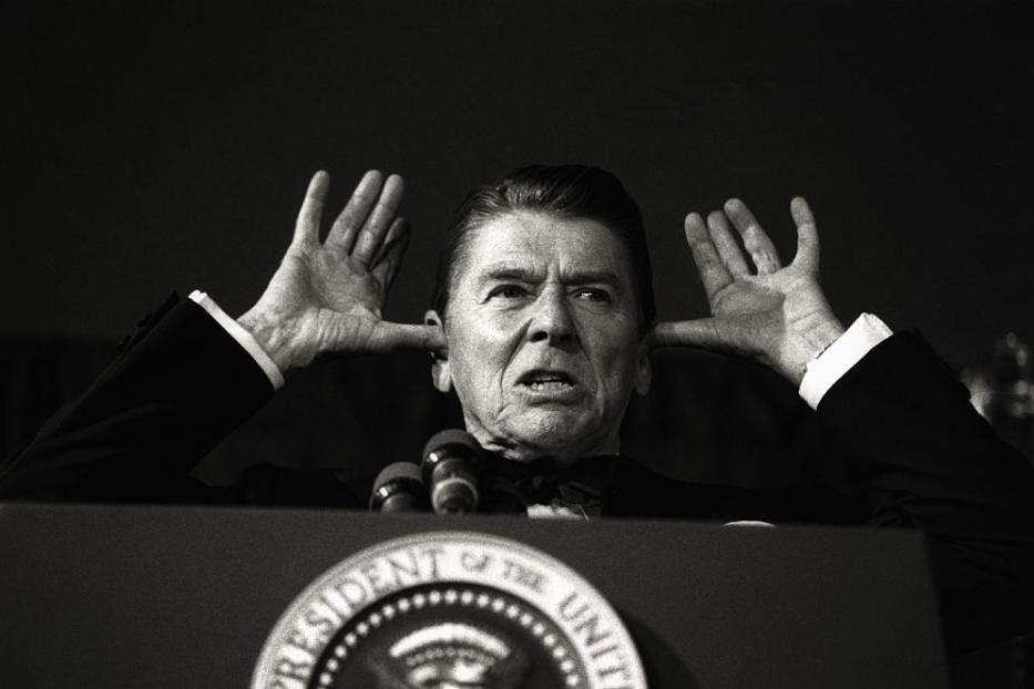 Ronald Reagan hält sich die Ohren zu, während er an einem Rednerpult steht.