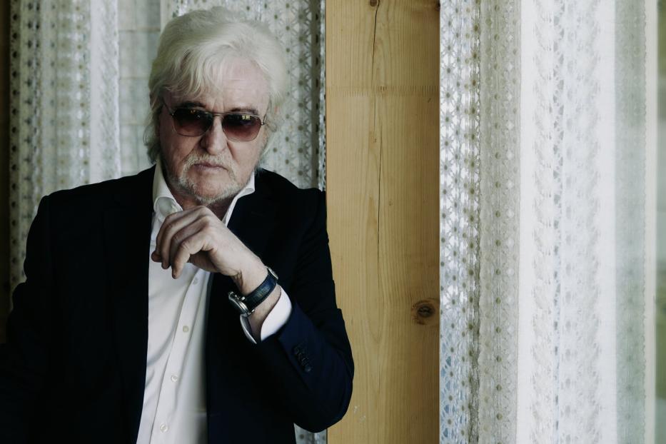 Ein älterer Mann mit Sonnenbrille und Anzug posiert vor einem Fenster.