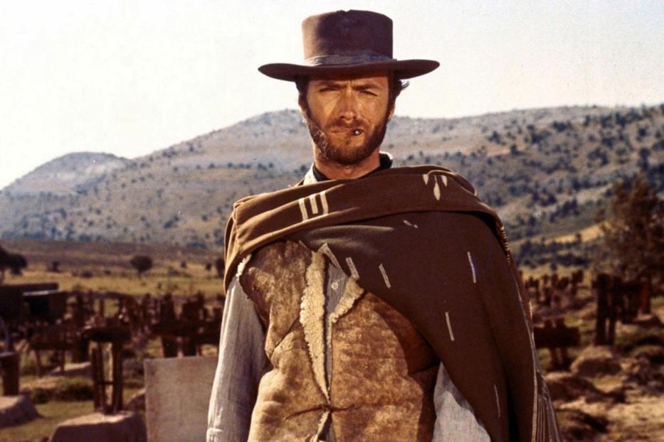 Clint Eastwood als „Blondie“ vor einer Hügellandschaft mit einem Friedhof im Hintergrund.
