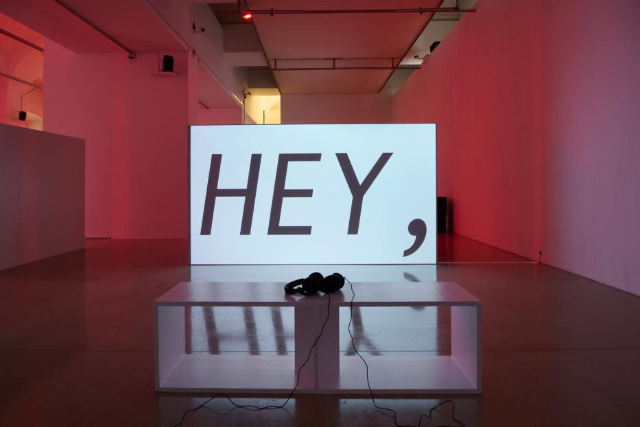 In einem Ausstellungsraum steht eine Leinwand mit dem Wort „HEY,“ darauf. Davor liegen Kopfhörer auf einem weißen Podest.