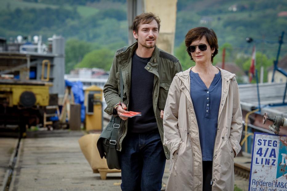 Ein Mann mit Notizbuch und eine Frau mit Sonnenbrille gehen an einem Kai entlang.