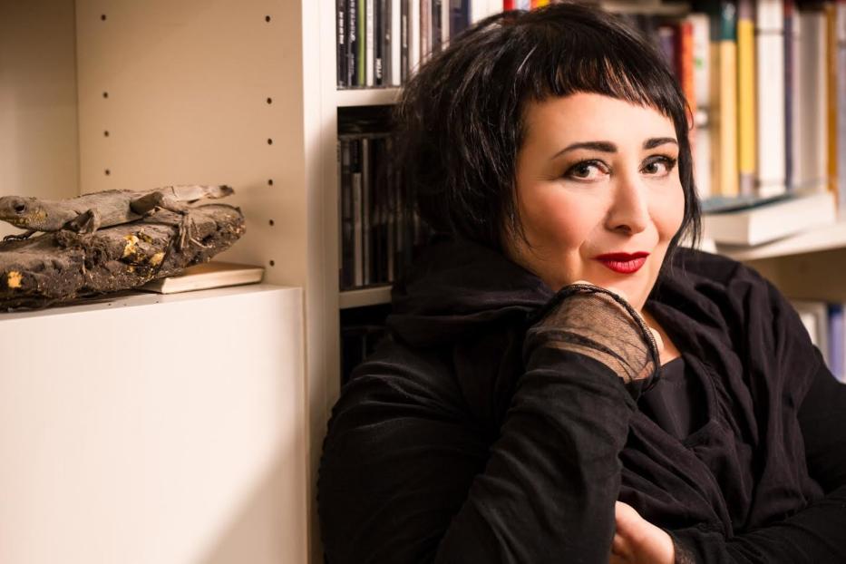 Eine Frau mit dunklen Haaren posiert vor einem Bücherregal mit einer Echse.