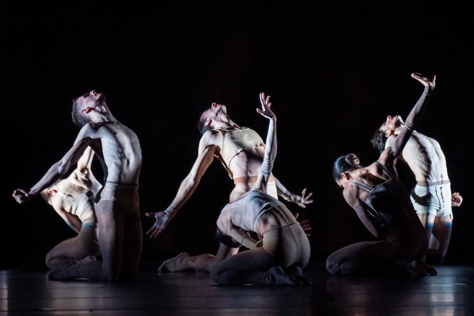 Fünf Tänzerinnen und Tänzer in beigen Kostümen führen eine synchrone Choreografie auf einer dunklen Bühne auf.