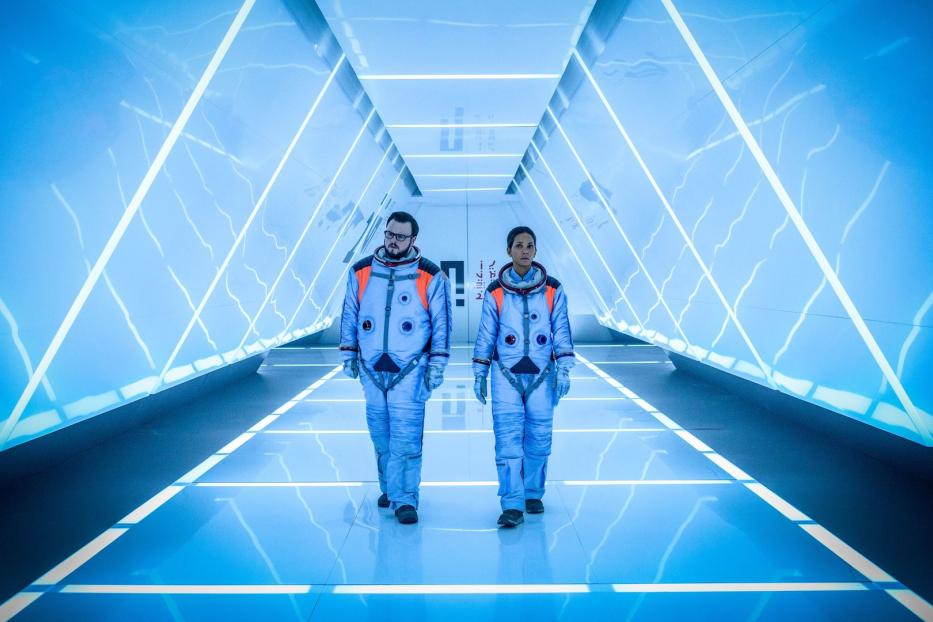 Zwei Astronauten gehen in Raumanzügen einen hell erleuchteten, futuristischen Korridor entlang.