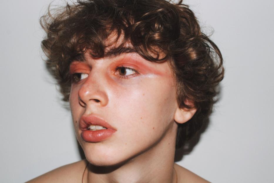 Ein junger Mann mit lockigem Haar und auffälligem, orangefarbenem Make-up blickt zur Seite.