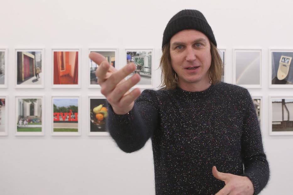 Ein Mann mit Mütze gestikuliert vor einer Wand mit gerahmten Fotos.