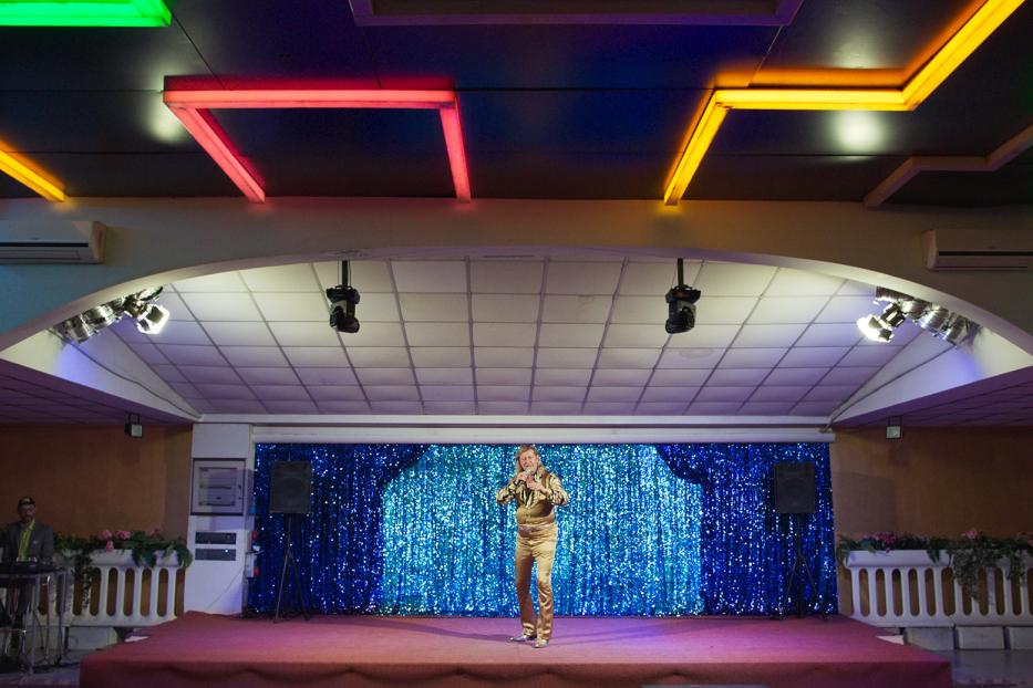 Ein Mann in einem goldenen Anzug steht auf einer Bühne mit blauem Glitzerhintergrund.