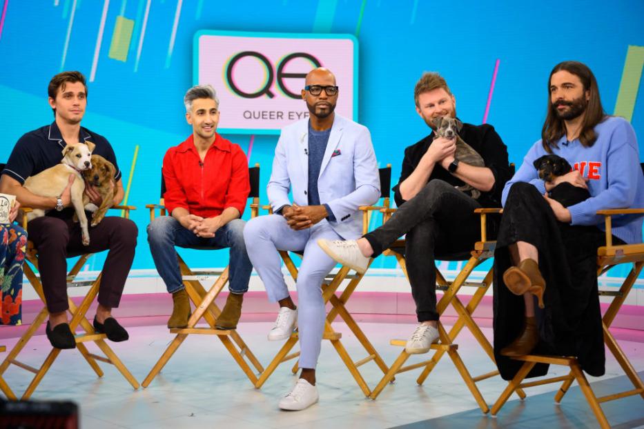 Die Besetzung von „Queer Eye“ sitzt mit Welpen auf Stühlen vor einem Queer Eye-Logo.