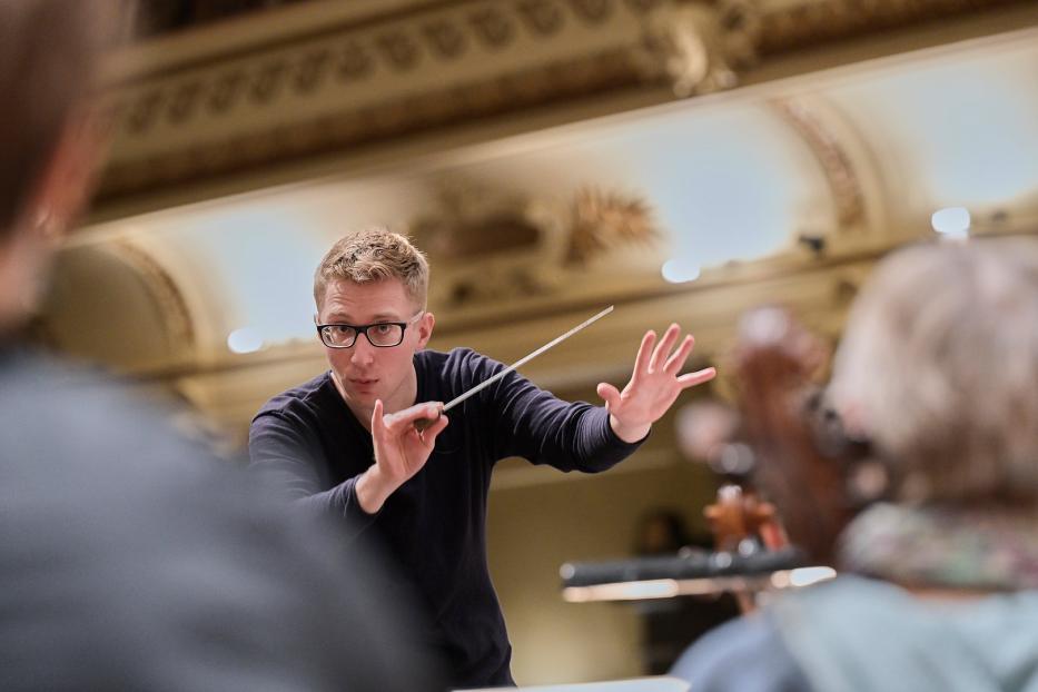 Ein Dirigent mit Brille dirigiert ein Orchester.