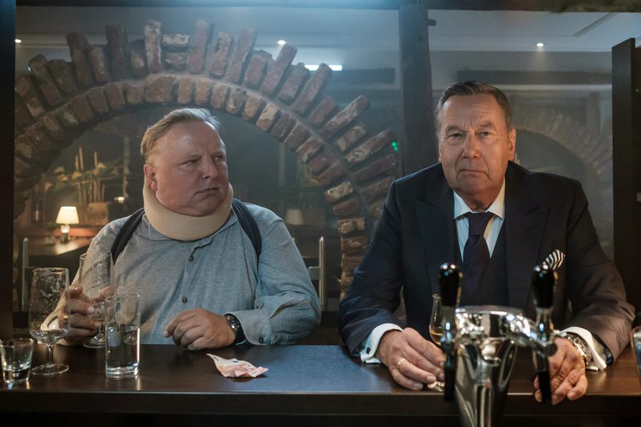 Zwei Männer sitzen an einer Bar, einer trägt eine Halskrause.