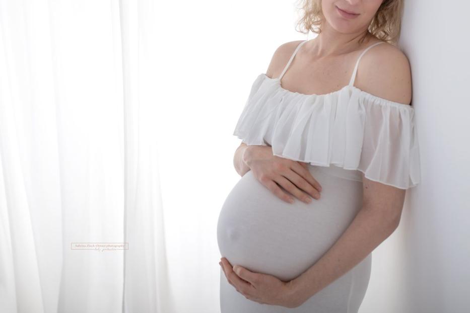 Eine schwangere Frau in einem weißen Kleid hält ihre Hände auf ihren Bauch.