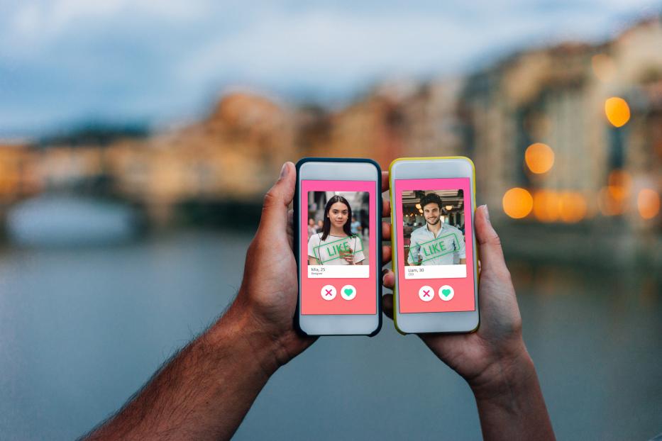 Eine Person hält zwei Smartphones mit Dating-App-Profilen vor einer Brücke.