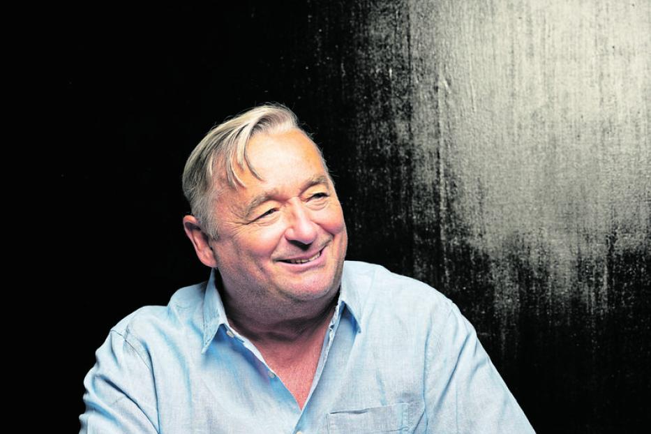 Ein lächelnder Mann mit hellblauem Hemd sitzt vor einem dunklen Hintergrund.