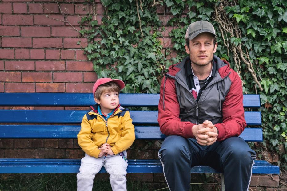Ein Mann und ein kleiner Junge sitzen auf einer blauen Bank vor einer Ziegelmauer mit Efeu.