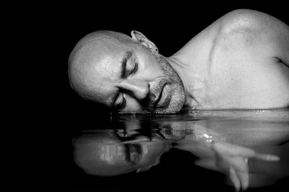 Ein Mann liegt mit geschlossenen Augen im Wasser, sein Spiegelbild ist sichtbar.