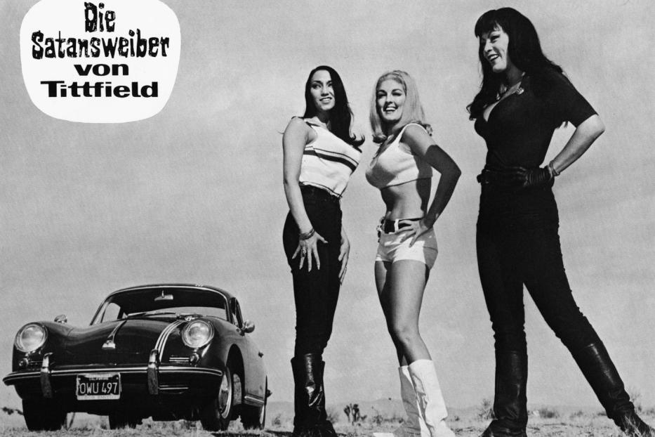 Drei Frauen posieren vor einem Porsche 356, im Hintergrund das Filmposter „Die Satansweiber von Tittfield“.