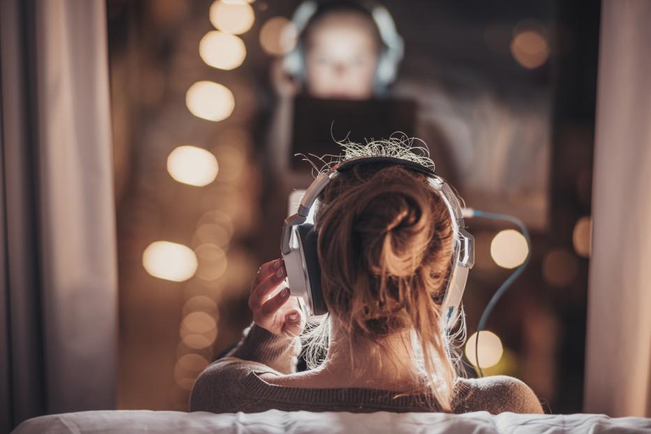 Eine junge Frau hört Musik über Kopfhörer, während eine weitere Person im Hintergrund ein Tablet benutzt.
