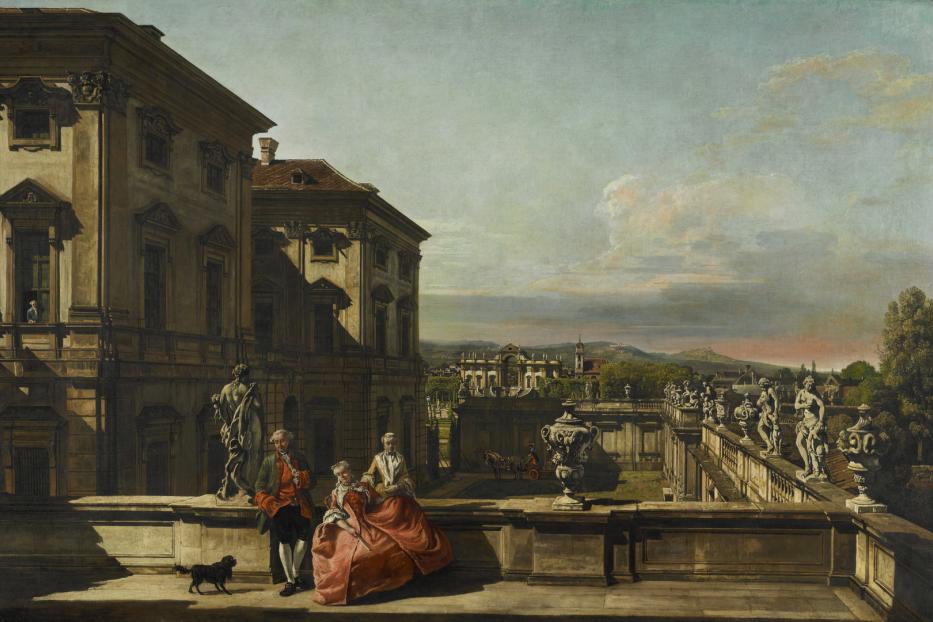 Ein Paar mit Hund auf einer Terrasse mit Blick auf ein Schloss und eine hügelige Landschaft.