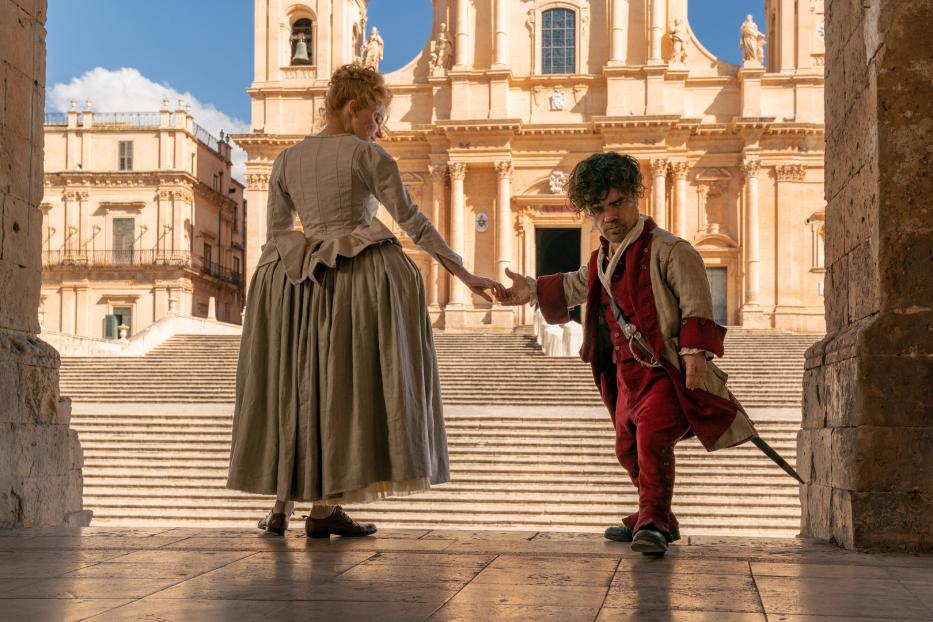 Peter Dinklage und eine Frau in historischer Kleidung stehen vor einem großen Gebäude.